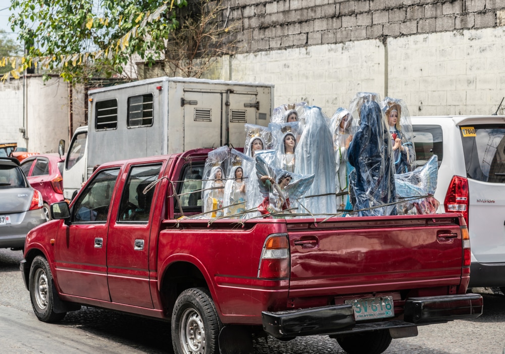 Maroon pickup truck with loading trunk full of small and larger Virgin Mary statues in street surrounded by traffic.
